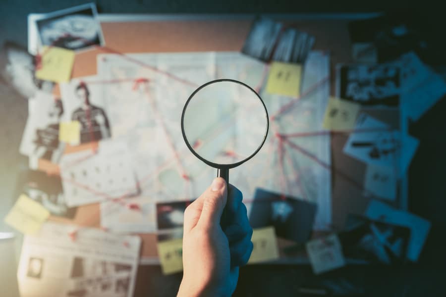 Detective Hand Holding A Magnifying Glass In Front Of A Board With Evidence, Crime Scene Photos And Map. High Contrast Image