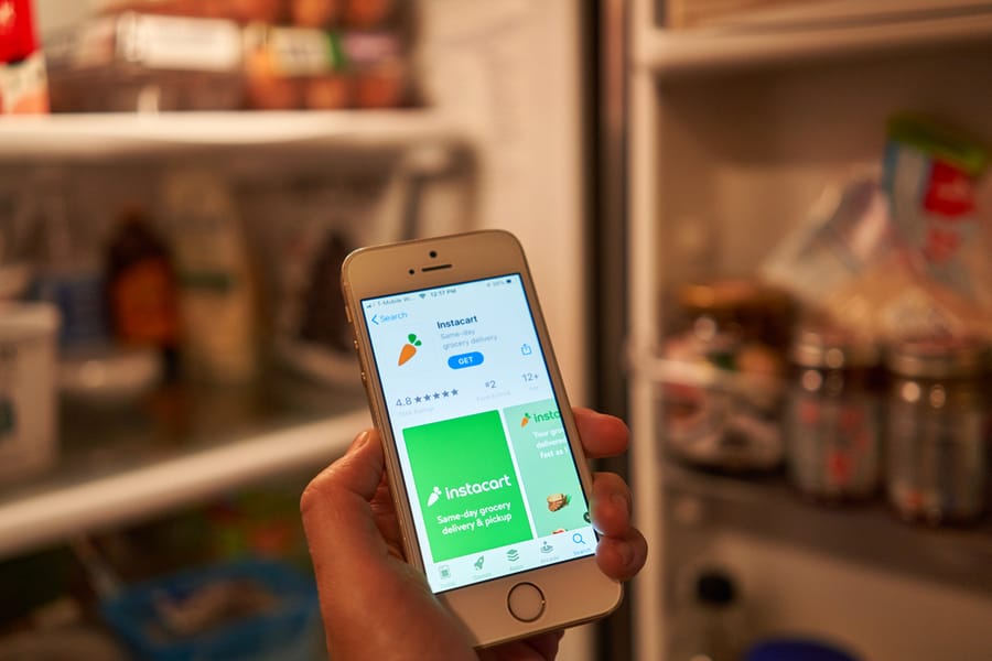 A Woman Checks The Instacart Mobile App On Her Phone While Opening The Refrigerator. Instacart Offers Same-Day Grocery Delivery And Pickup Service In The Us &Amp; Canada.