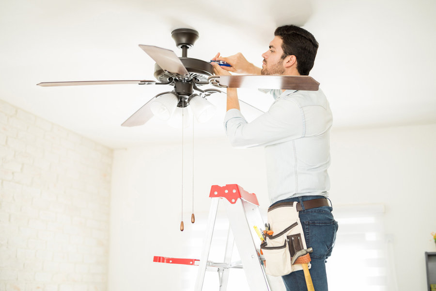 Attractive Young Handyman Stepping On A Ladder And Fixing A Ceiling Fan