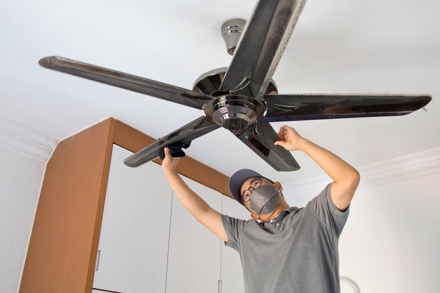Low Angle View Of An Asian Man Cleaning Ceiling Fan At Home.