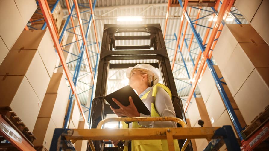 Professional Female Worker Wearing Hard Hat Lifts Herself On Aerial Work Platform To Check Stock And Inventory With Digital Tablet On The Higher Level Of Retail Warehouse Full Of Shelves With Goods.